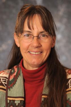 Image of a head shot of a woman wearing a native american patterned jacket in front of a gray backgound